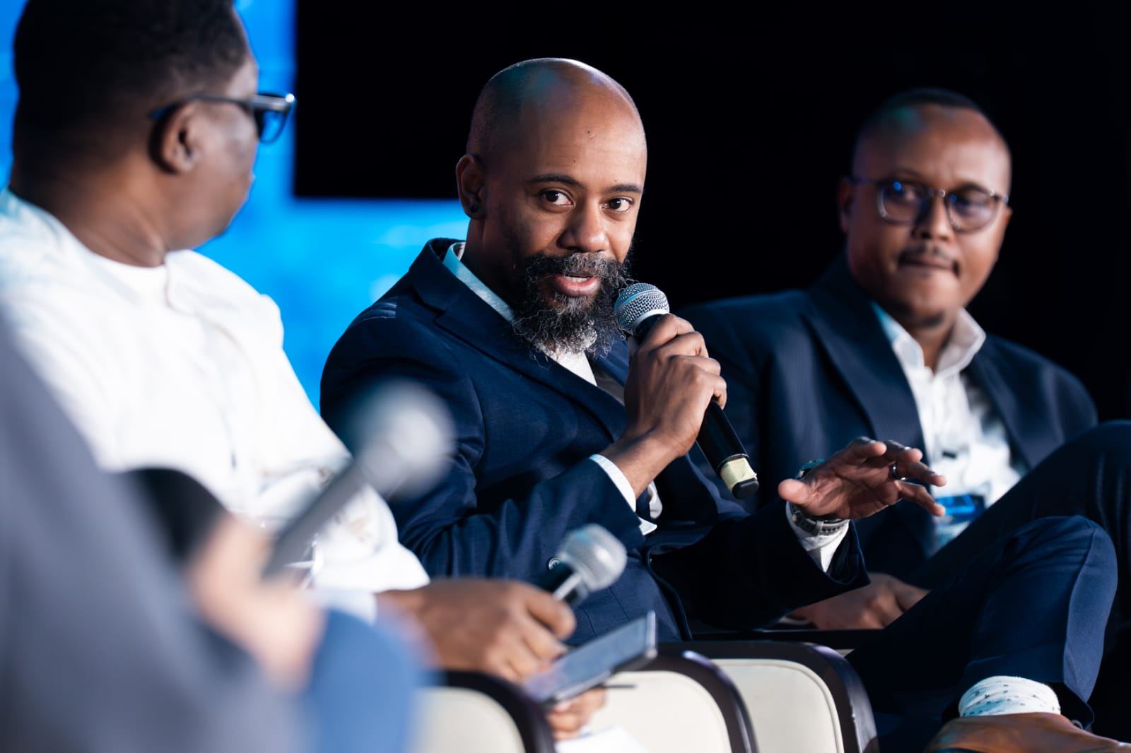 A panel discussion in progress, three speakers seated, one of them speaking into a microphone.